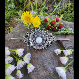 Full view of a crescent moon-shaped hair stick with a radiant opalescent stone centerpiece surrounded by intricate Victorian-style filigree, displayed on a textured stone surface surrounded by freshly picked wildflowers including dandelions, buttercups, and white blossoms