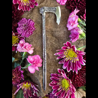 Viking hatchet hair stick placed on a rocky surface surrounded by vibrant pink and purple flowers for an enchanting aesthetic